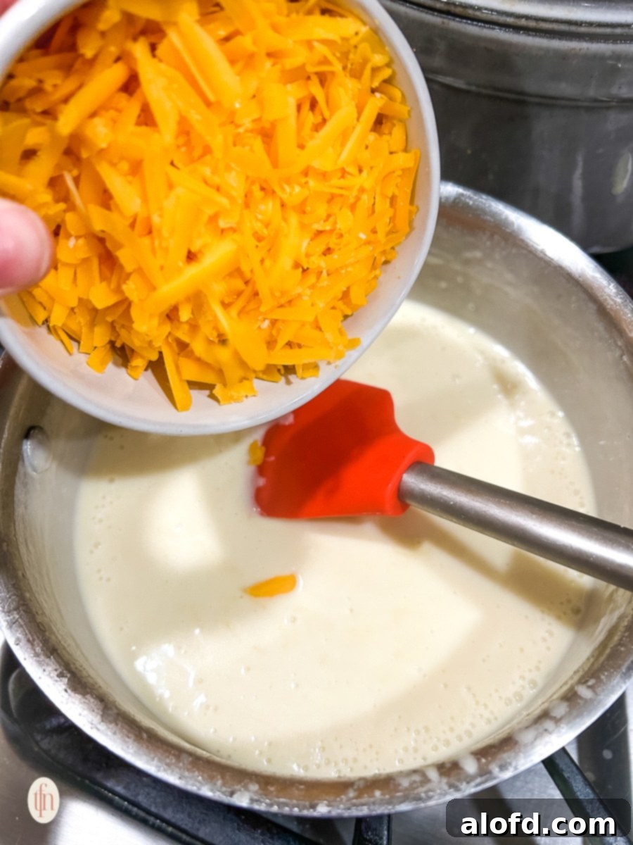 A hand gently adding a bowl of freshly shredded cheese into a creamy béchamel sauce, ready to be melted and mixed.