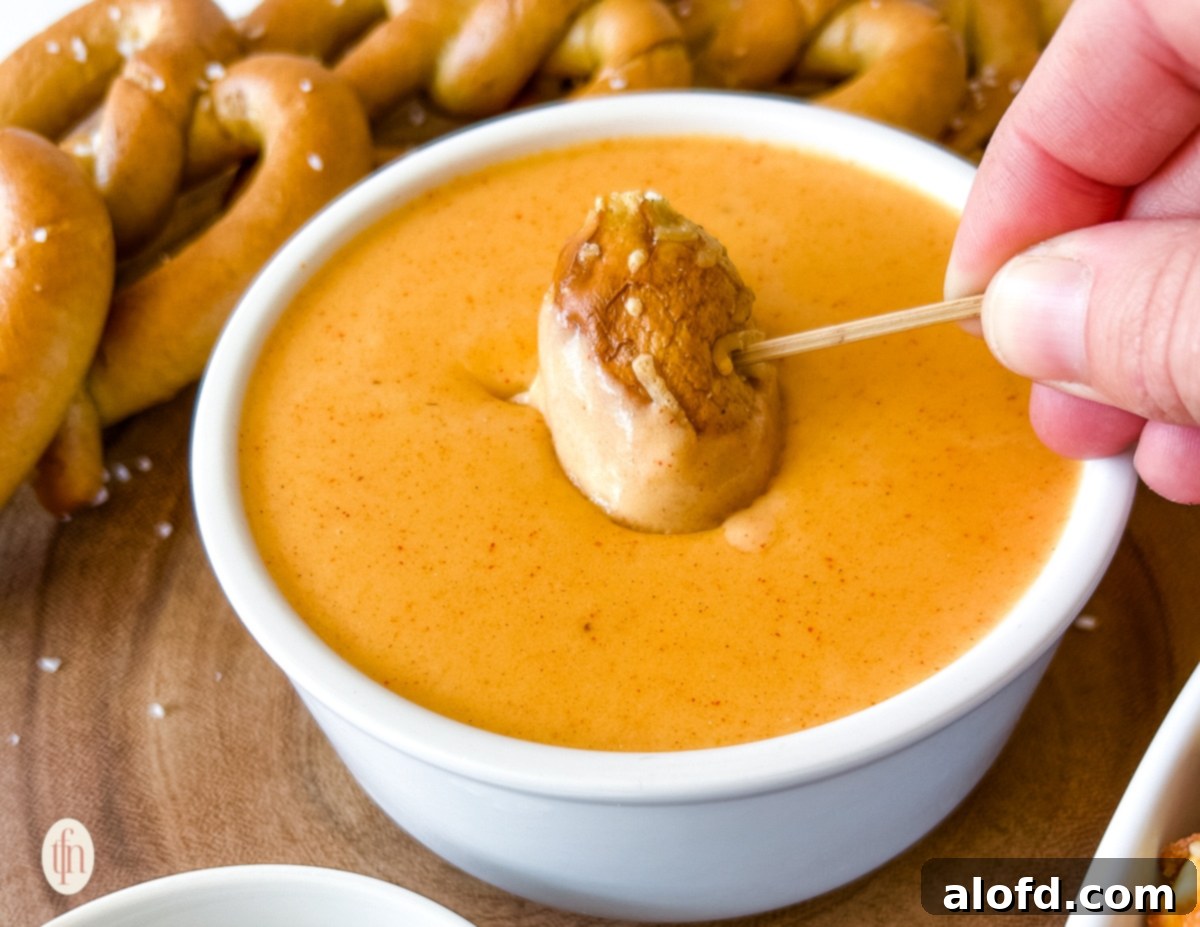 A close-up of a hand dipping a piece of soft pretzel into a bowl of creamy pretzel cheese dip, highlighting its luscious texture.