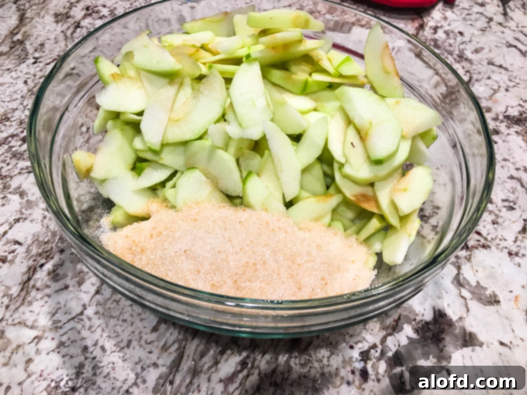 Freshly peeled, cored, and thinly sliced apples tossed with granulated sugar in a mixing bowl, ready for the baking dish.