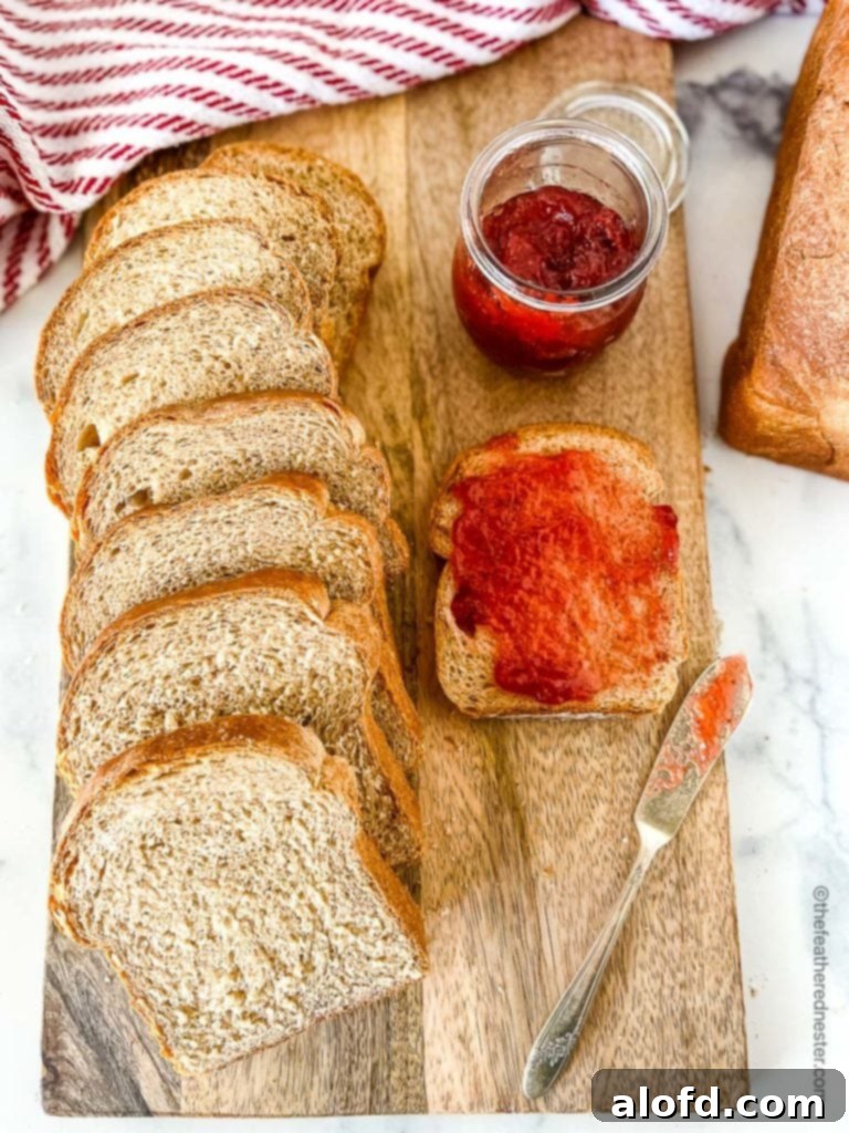 A beautifully sliced loaf of homemade whole wheat sandwich bread on a wooden board, with a slice spread with strawberry jam next to a jar of jam, highlighting the freshness and deliciousness.