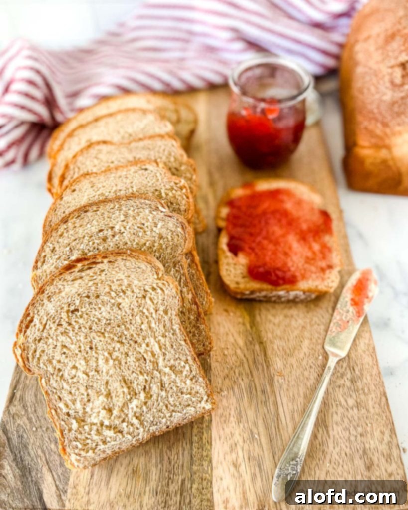 A wooden board displays a freshly baked loaf of sliced whole wheat bread, with a single slice generously spread with strawberry jam next to an open jam jar, highlighting its homemade goodness.