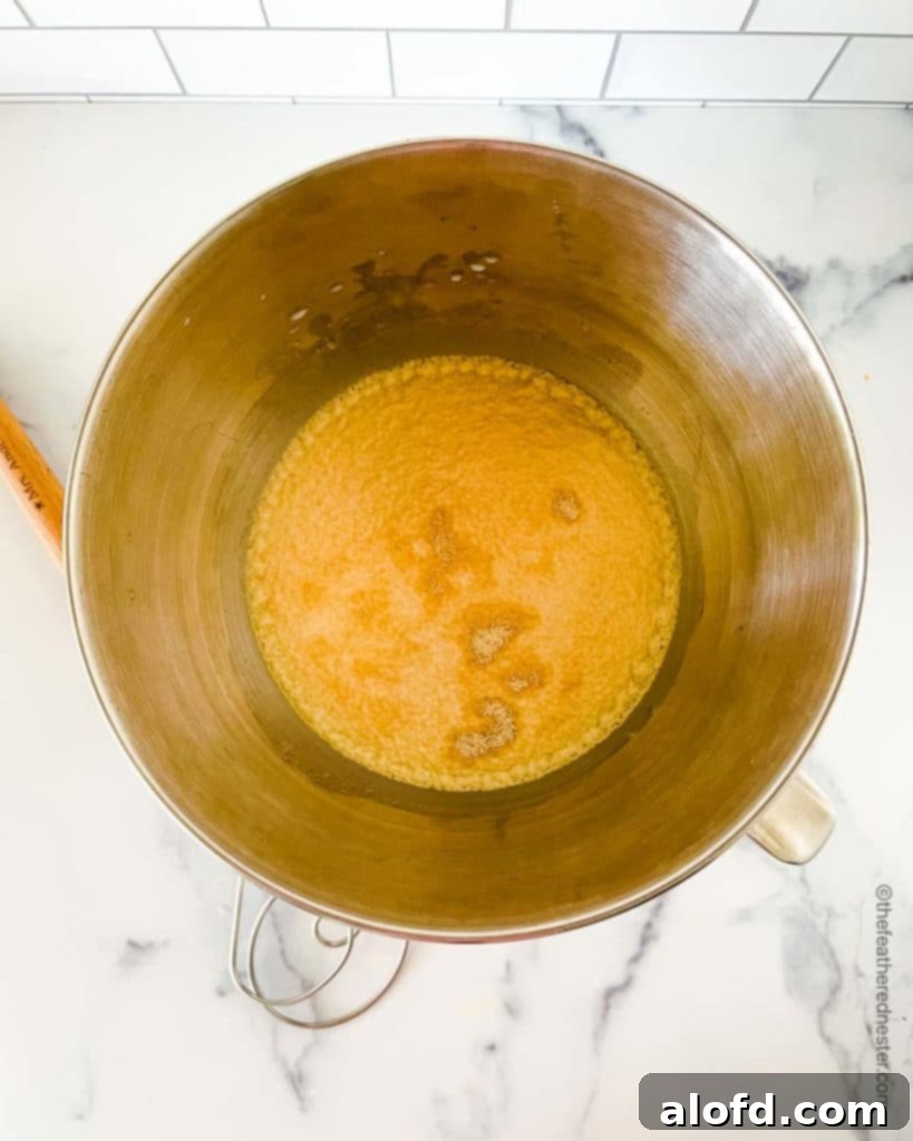 A stainless steel mixing bowl shows milk, water, and sugar with active dry yeast sprinkled on top, beginning the activation process for making delicious wheat sandwich bread.