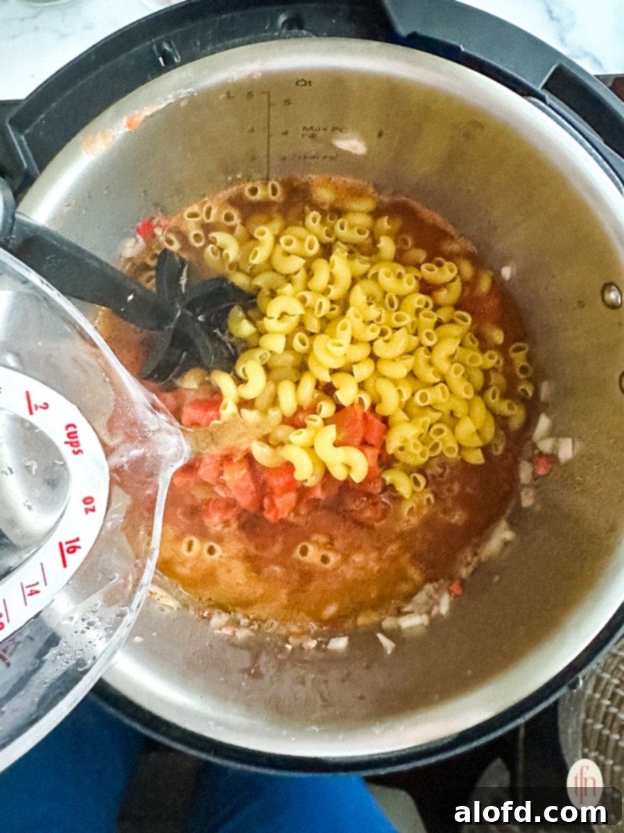 Adding hot water to a pot containing elbow macaroni and other chili ingredients, preparing for pressure cooking.