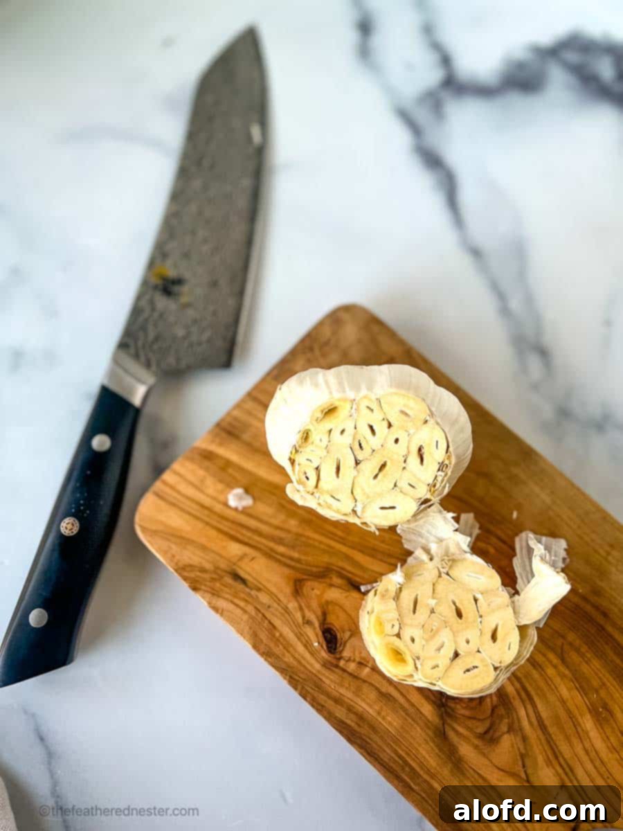 A whole head of garlic with the top sliced off, ready for roasting, positioned next to a chef's knife on a clean white surface.