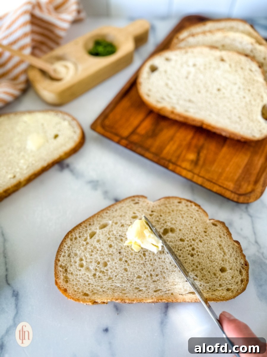 A hand gently spreading softened butter onto a thick slice of homemade sourdough garlic bread with a butter knife, ready for toasting.