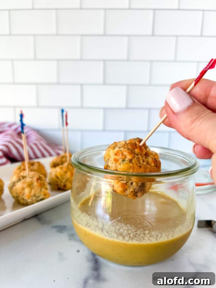 A pimento cheese sausage ball being dipped into spicy mustard sauce, with a tray of more sausage balls in the background.