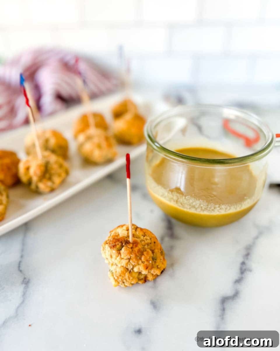 An appetizer toothpick stuck in a pimento cheese sausage ball, with dipping sauce and a tray of more appetizer balls in the background.