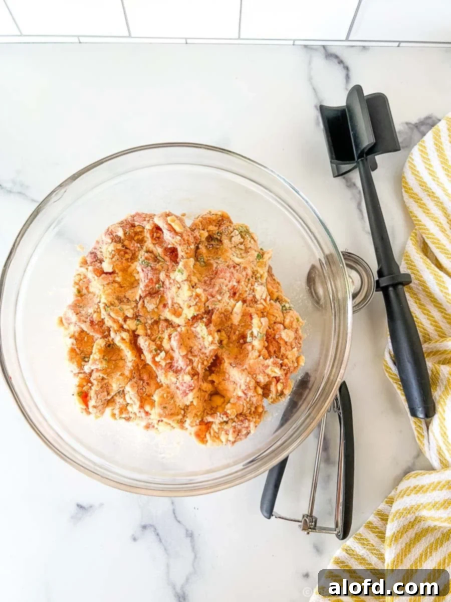 A clear bowl of mixed sausage ball ingredients including ground sausage, pimento cheese, and baking mix, with a scoop and meat chopper nearby.