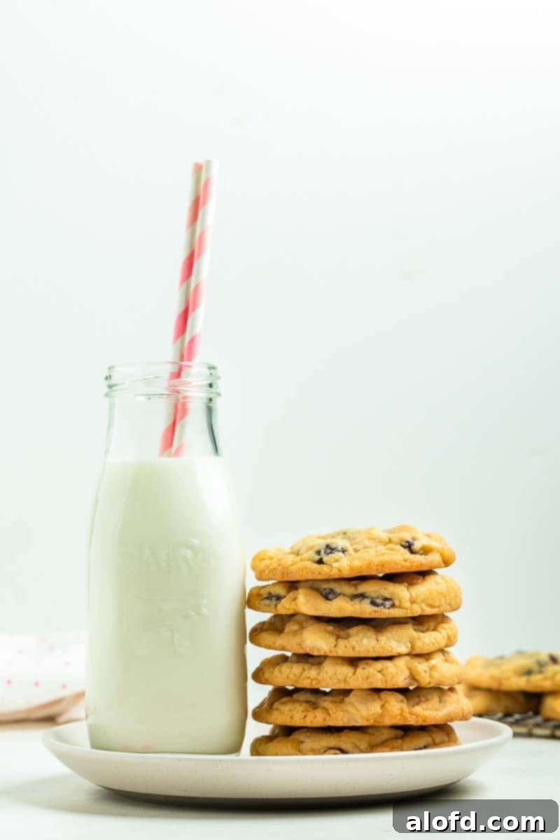 A bottle of milk with a straw beside a stack of perfectly baked white chocolate raspberry cookies on a pristine white plate, inviting you for a delicious treat.