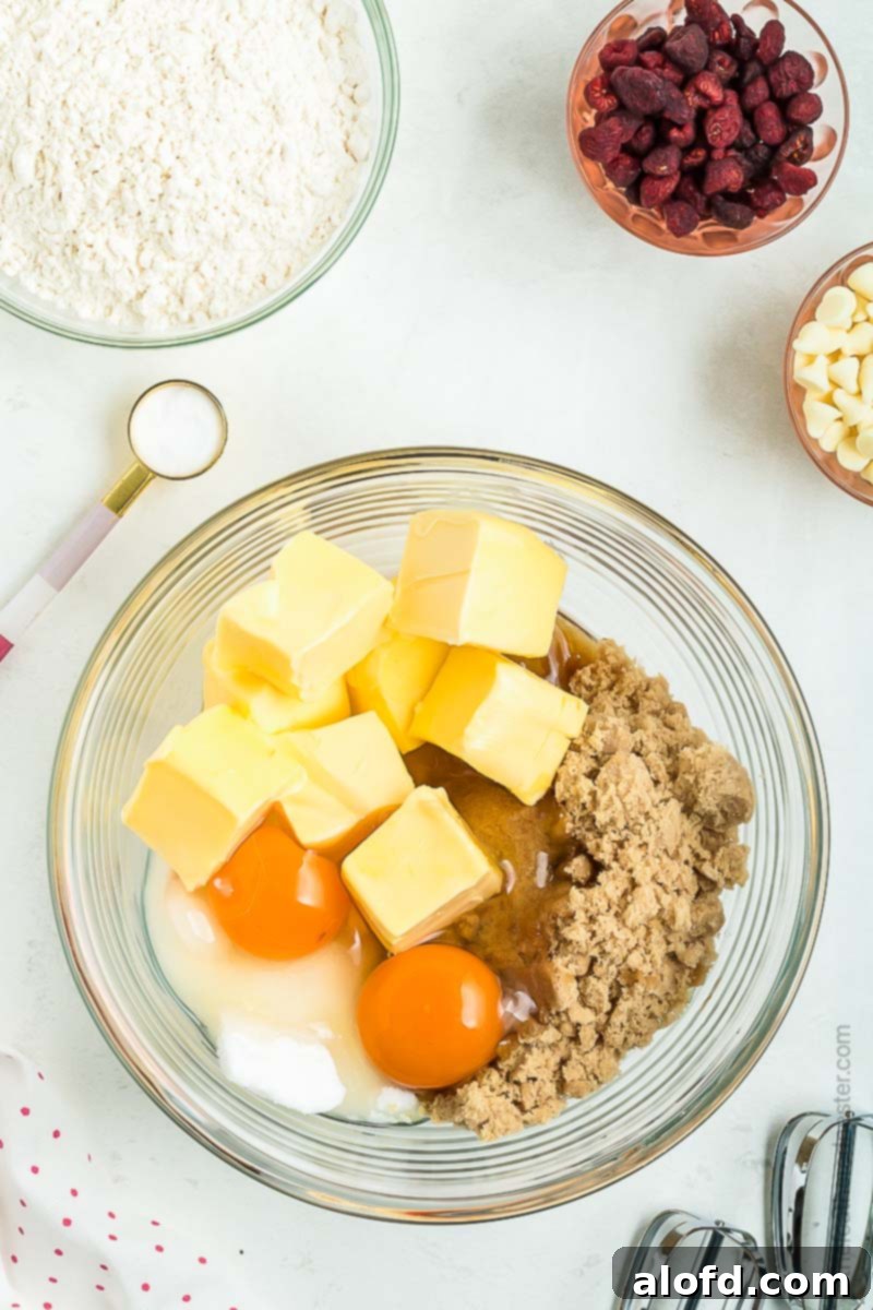 A bowl containing a mixture of sugars, butter, and eggs is surrounded by smaller bowls holding flour, dried raspberries, and white chocolate chips, illustrating the ingredients ready for mixing.