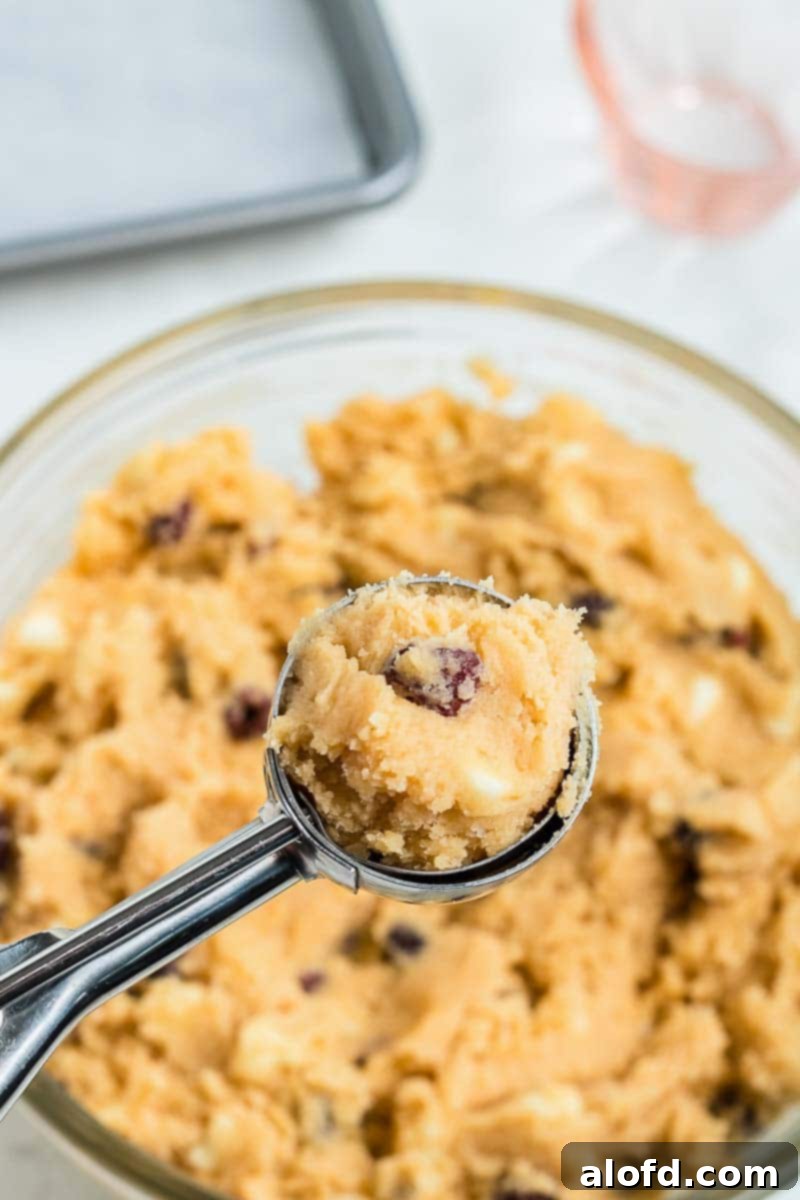 A perfectly portioned scoop of cookie dough rests on a baking sheet, with the mixing bowl filled with more dough blurred in the background, showcasing the next step in the baking process.