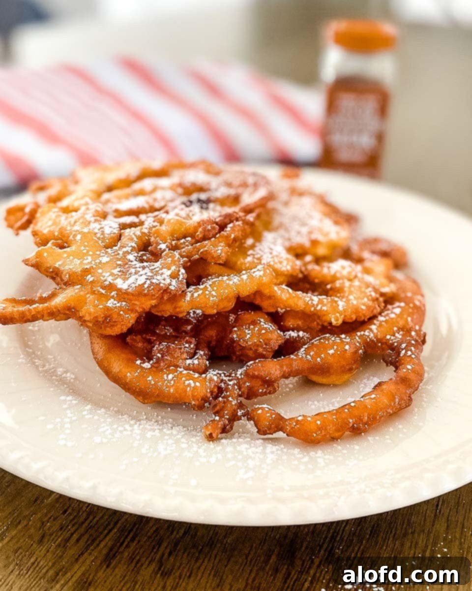 A white plate featuring a perfectly fried Bisquick funnel cake, garnished with powdered sugar. A red striped cloth and a cinnamon powder bottle are visible in the background, hinting at classic toppings.