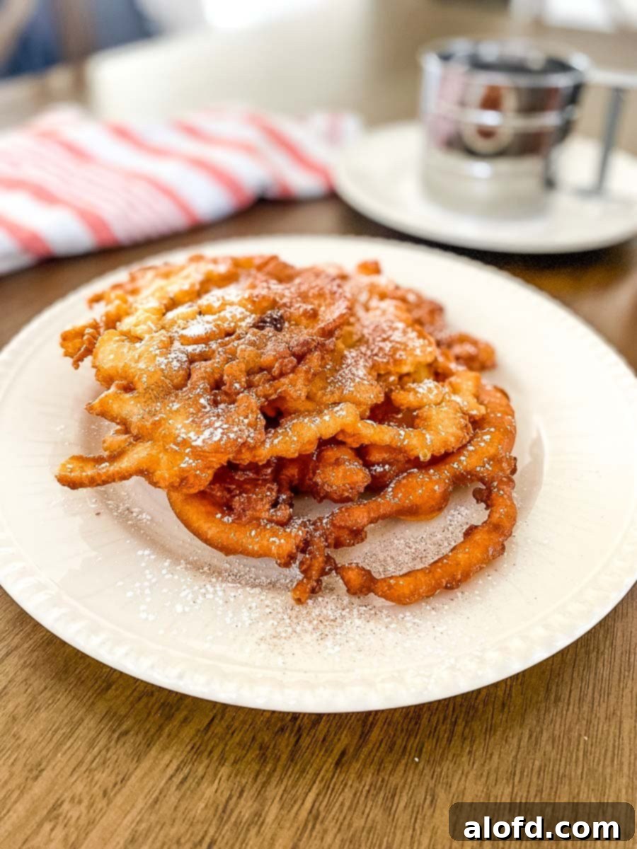 A beautifully presented homemade funnel cake on a white plate, accompanied by a striped red cloth and a silver cup in the background, ready for a delightful treat.