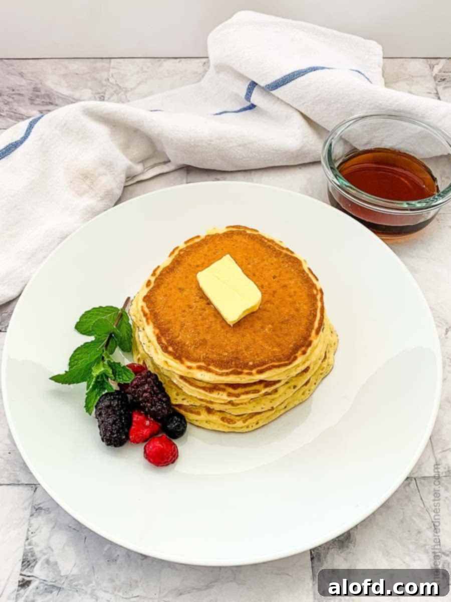 Overhead view of a stack of fluffy pancakes garnished with berries and a pat of butter, with a bottle of syrup in the background.