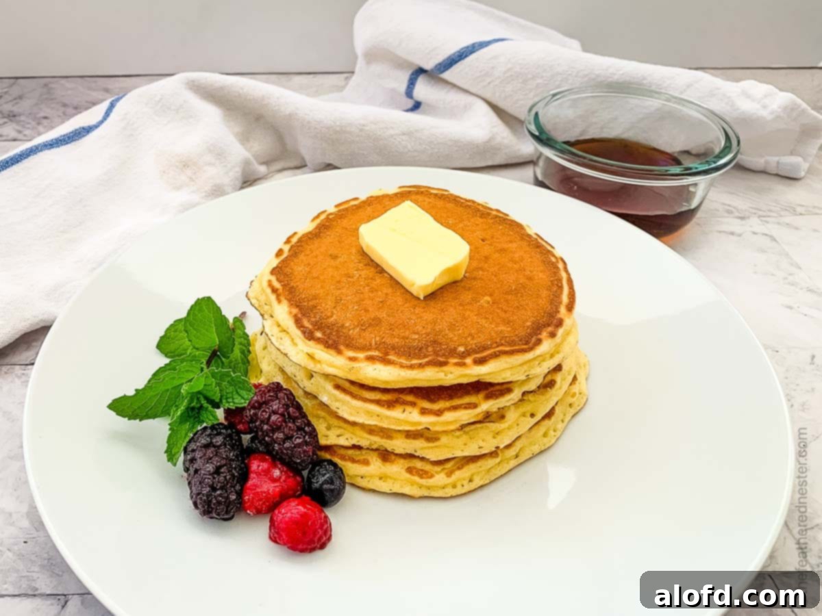 A white plate of pancakes garnished with fresh berries and butter, with a container of syrup in the background.