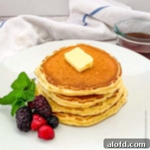A white plate of pancakes garnished with berries and butter, topped with a container of syrup in the background.