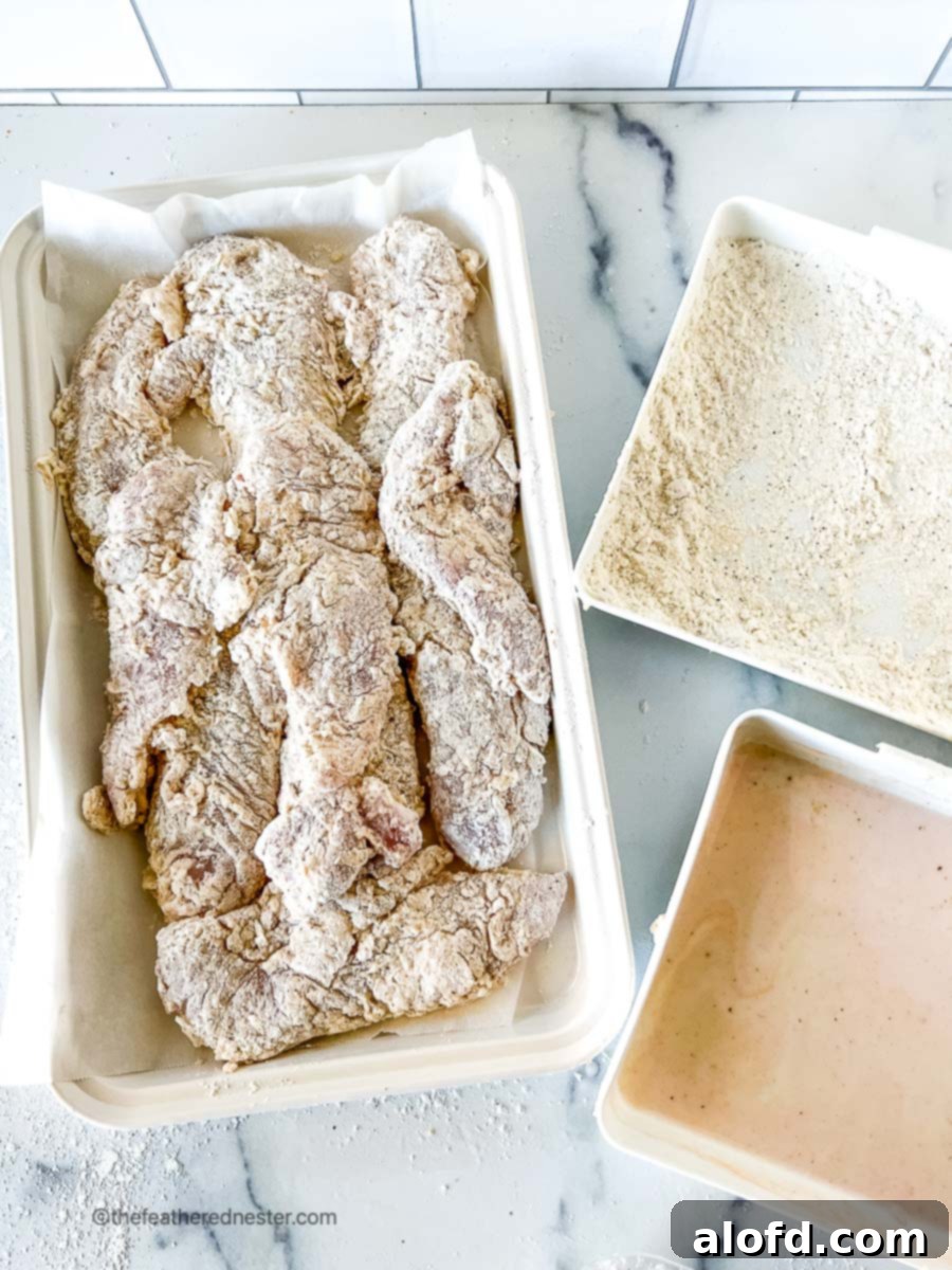 Fiery Nashville Chicken Strips 6 Chicken tenders that are coated in flour and buttermilk mixture on a white serving platter along with flour mixture and buttermilk mixture on separate trays.