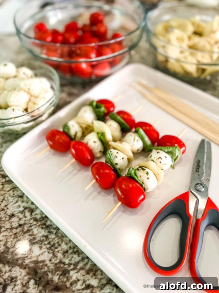 Caprese Tortellini Poppers 8 Bowls of cherry tomatoes, mozzarella balls, pasta, and basil leaves along with a tray of 4 assembled tortellini appetizers and a pair of scissors on a white serving tray