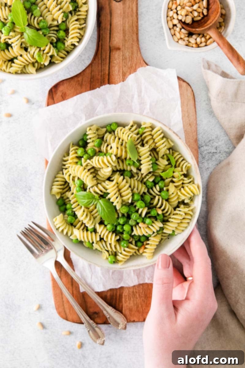 Spring Pea Pasta 11 A hand holding a plate of vibrant rotini pasta salad with green vegetables, with silverware neatly arranged beside it.
