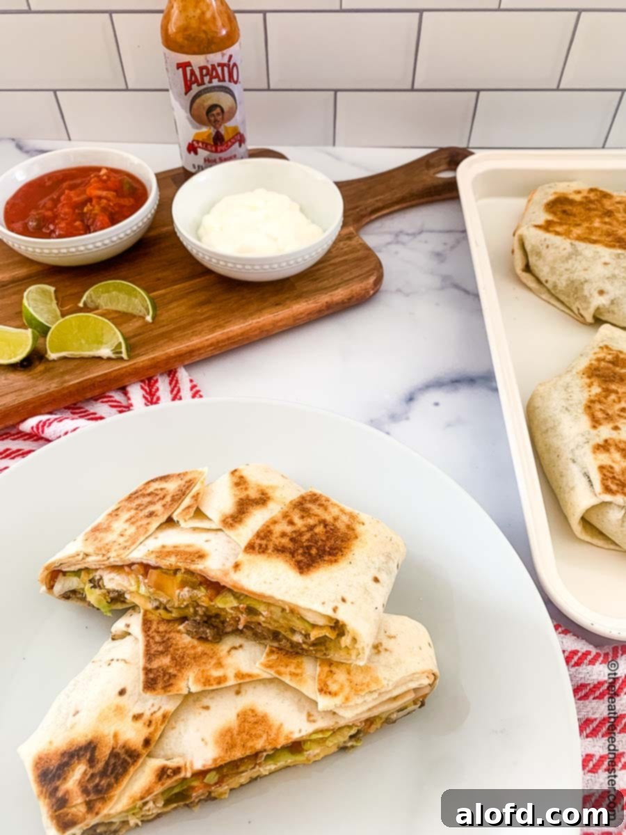 Blackstone's Ultimate Crunchwrap 2 Homemade crunchwrap on a white plate with a serving board featuring bowls of sour cream, salsa, a bottle of Tapatio, and lime slices in the background, ready to be enjoyed.