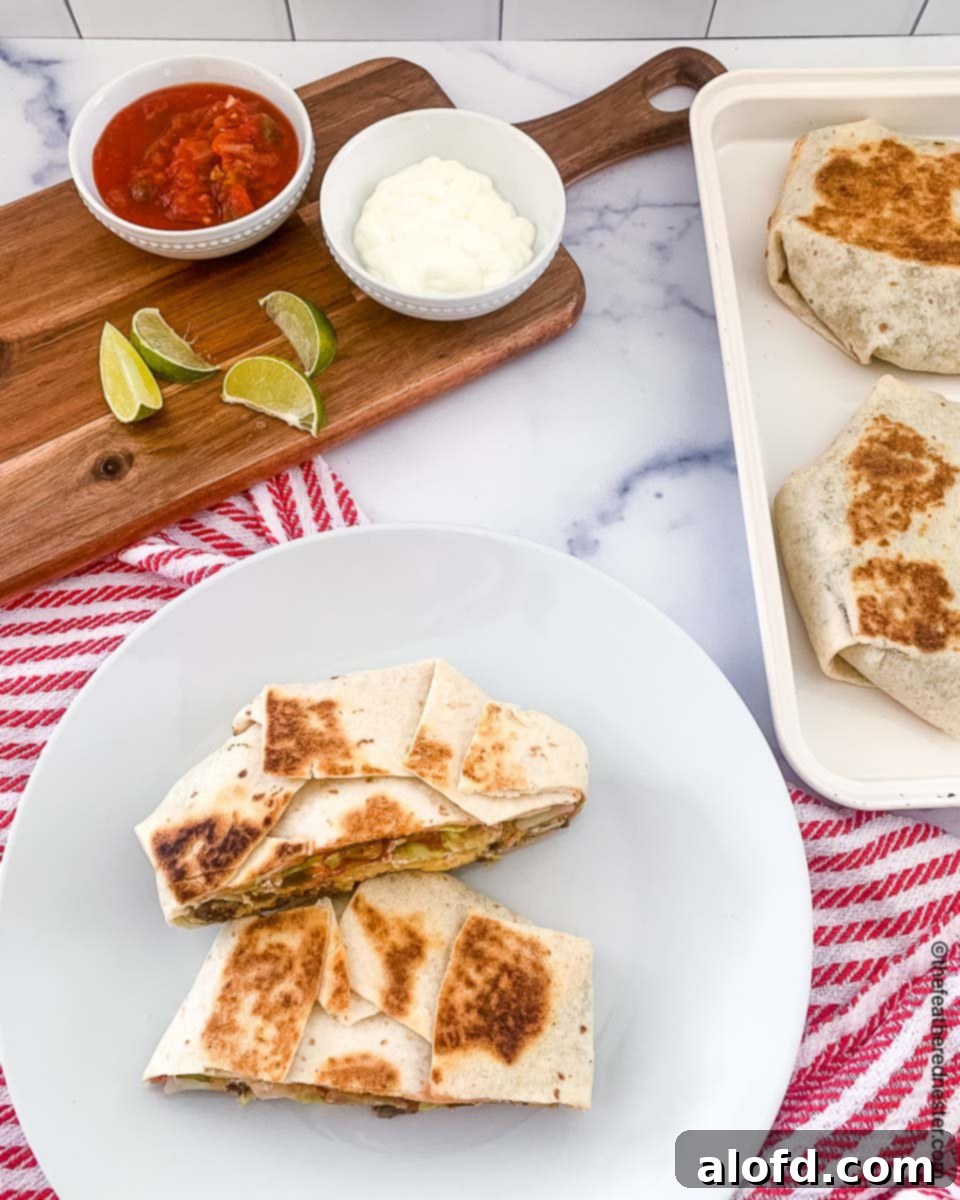 Blackstone's Ultimate Crunchwrap 3 A perfectly cooked crunchwrap cut in half, showcasing its layers, served on a white plate alongside another whole crunchwrap on a tray. In the background, bowls of sour cream, salsa, and lime slices are artfully arranged on a serving board.