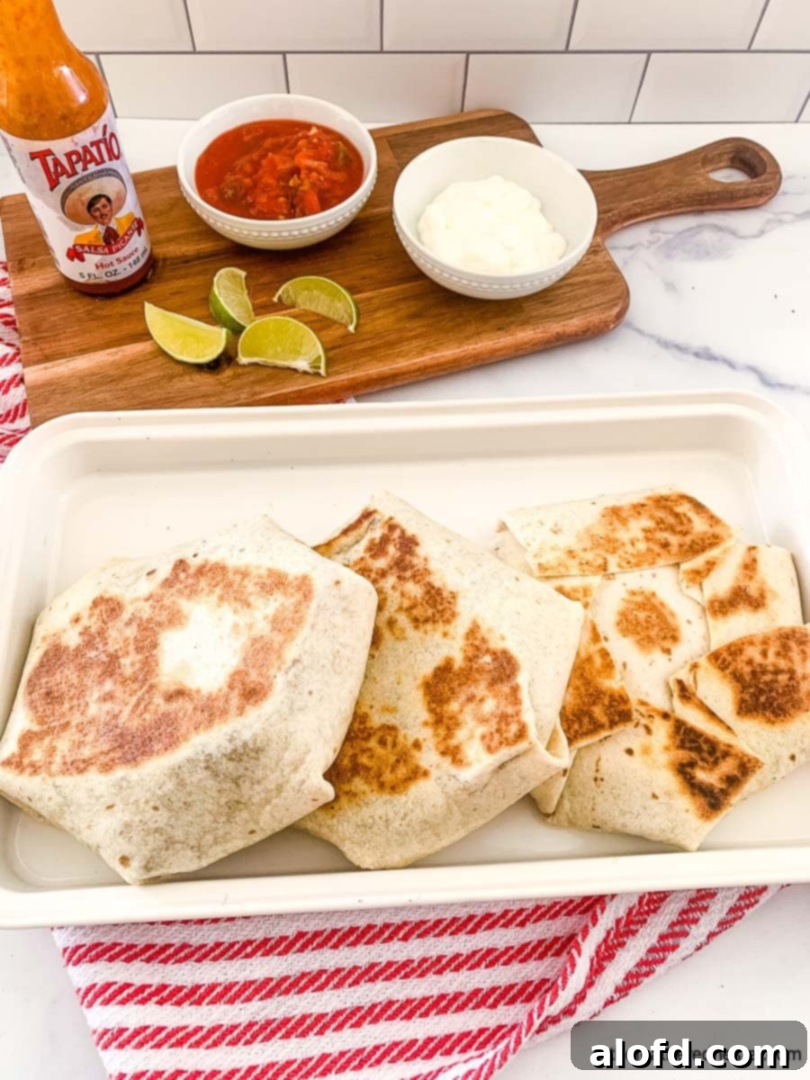Blackstone's Ultimate Crunchwrap 5 A finished Blackstone crunchwrap resting on a white plate, with a serving board in the background holding bowls of sour cream, salsa, a bottle of Tapatio hot sauce, and fresh lime slices, ready for a delicious meal.