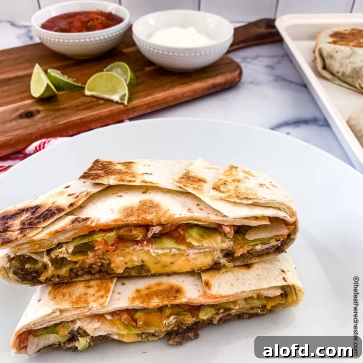 Blackstone's Ultimate Crunchwrap 8 A perfectly cooked copycat crunchwrap, sliced in half to reveal its layers, presented on a white plate. In the background, a serving board holds bowls of sour cream, salsa, a bottle of Tapatio, and fresh lime slices, ready for customization.