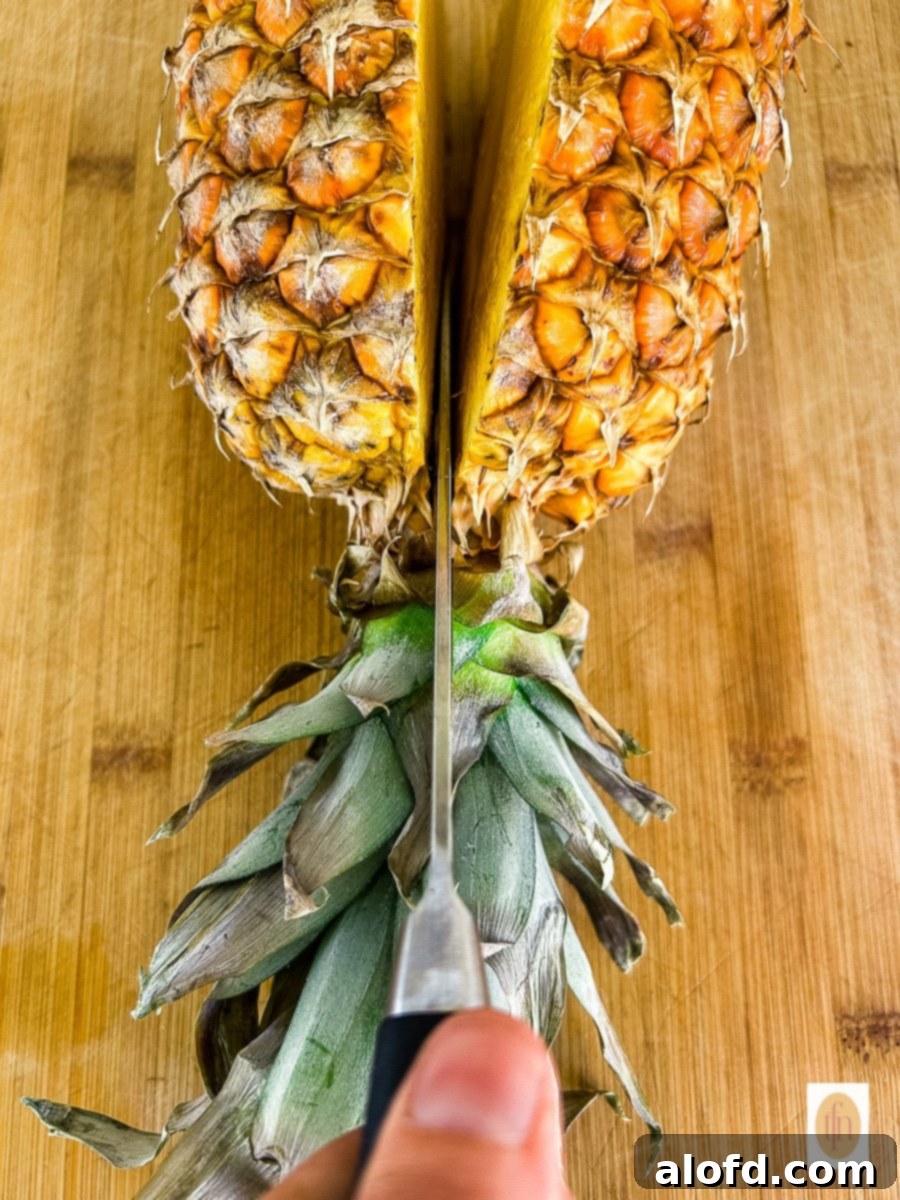 A large knife is used to cut a fresh pineapple in half, preparing it for the bowls.