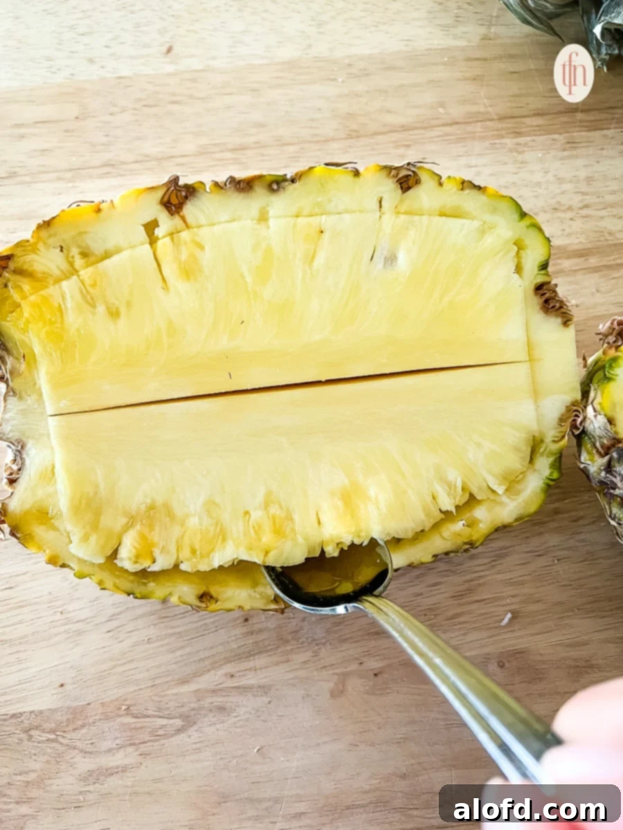 A spoon is used to carefully remove the fruit and fibrous core from a halved pineapple shell.