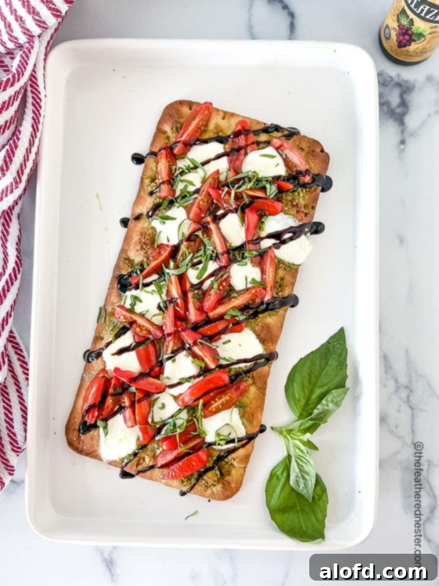 Classic Margherita Flatbread 2 A white serving tray with a freshly baked Caprese flatbread pizza garnished with a sprig of fresh basil, placed next to a red and white napkin.