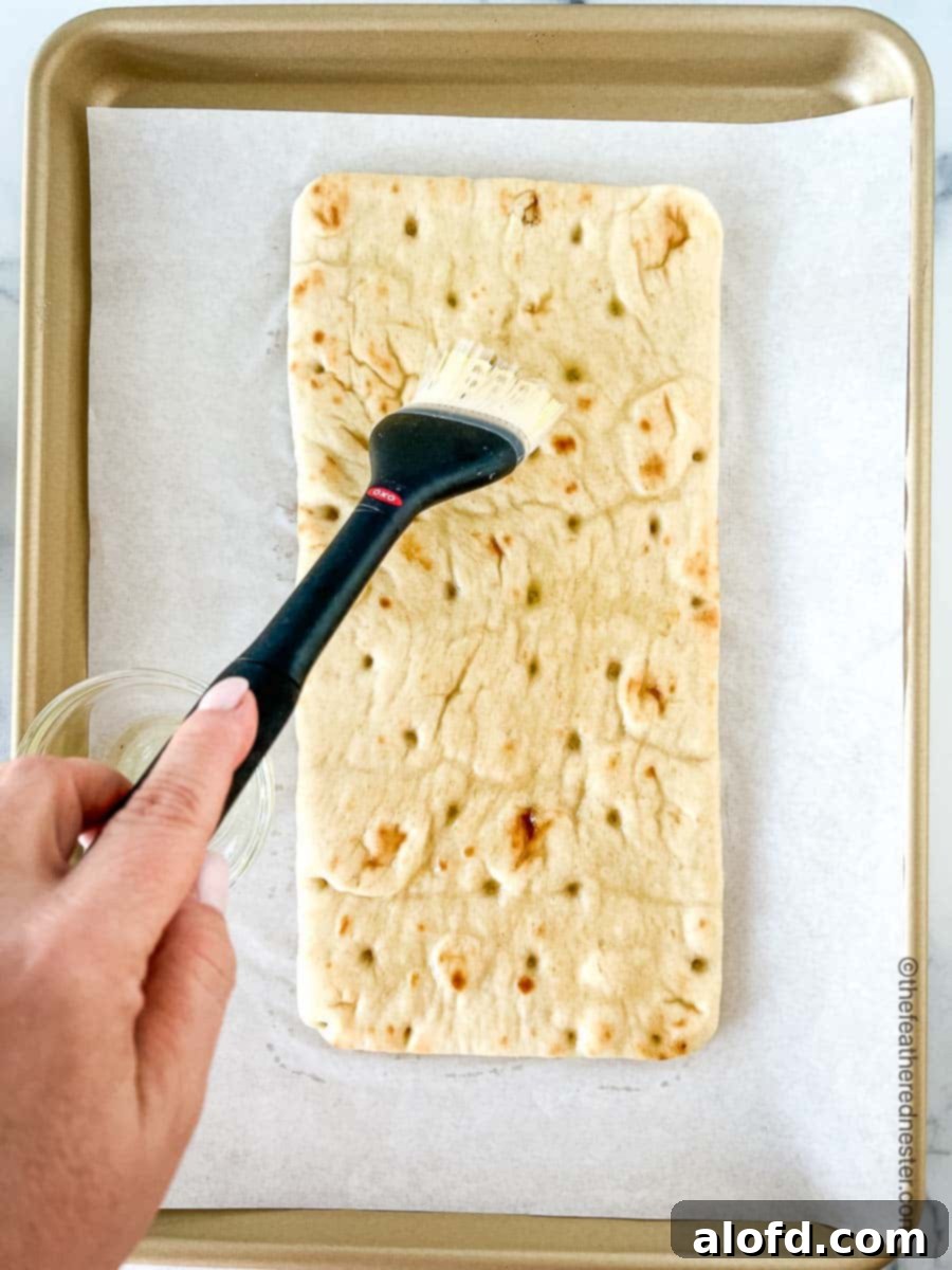 Classic Margherita Flatbread 6 A hand brushing olive oil onto a flatbread placed on a parchment-covered baking sheet.