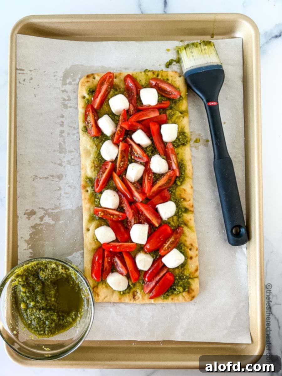 Classic Margherita Flatbread 7 A baking sheet featuring a bowl of fresh pesto sauce and a flatbread generously topped with pesto, halved grape tomatoes, and fresh mozzarella cheese.