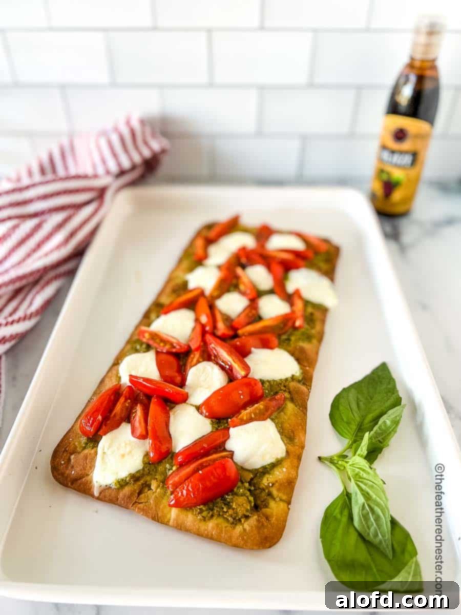 Classic Margherita Flatbread 8 A parchment-covered baking sheet with a baked flatbread topped with pesto, tomatoes, and mozzarella cheese, with a bottle of balsamic glaze and a red and white napkin nearby.