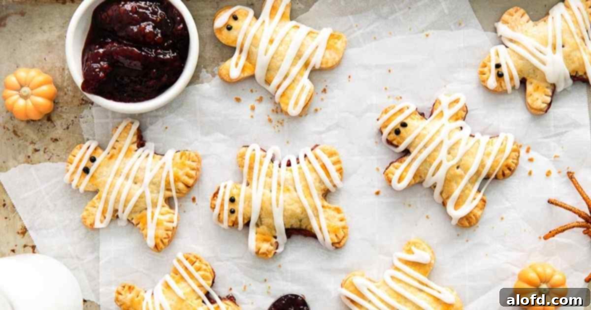 a baking sheet of halloween mummy hand pies with a white mini bowl of berry jam on top of parchment paper.