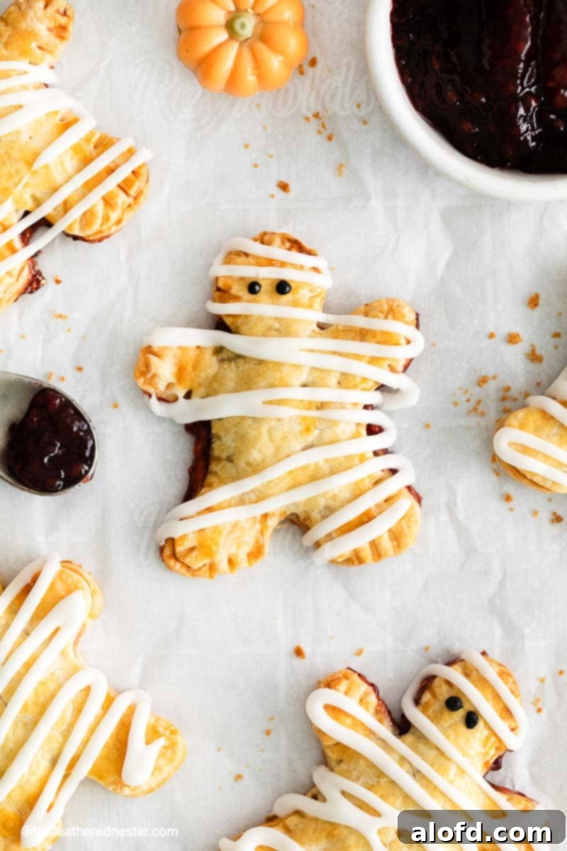 Halloween hand pie on top of a parchment paper with another mummy hand pies and a spoon and bowl of berry jam beside it.