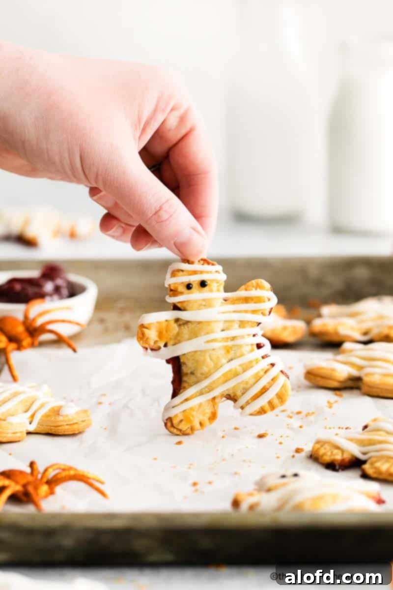 a hand holding a mummy hand pie placed on a parchment paper and baking sheet with spider toy, pumpkin toy and a bowl of berry filling at the background