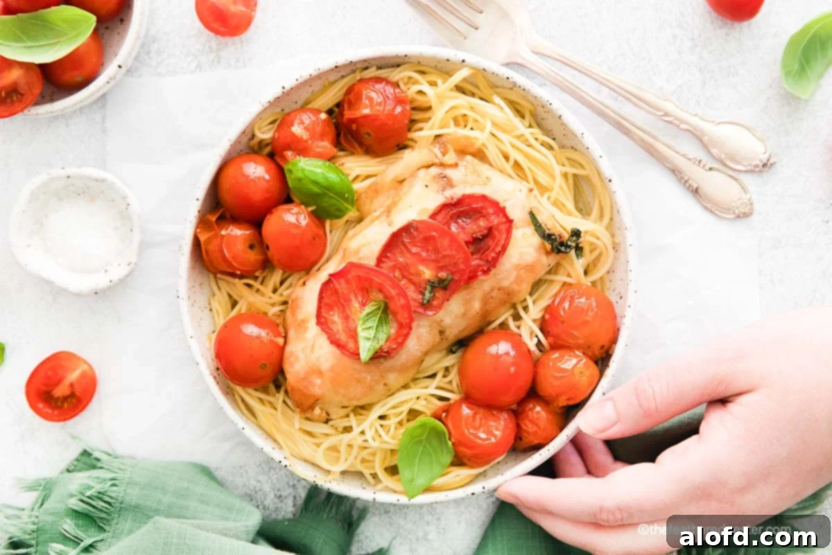 One Pan Caprese Chicken 11 A hand holding a white bowl of pasta and chicken with roasted tomatoes.