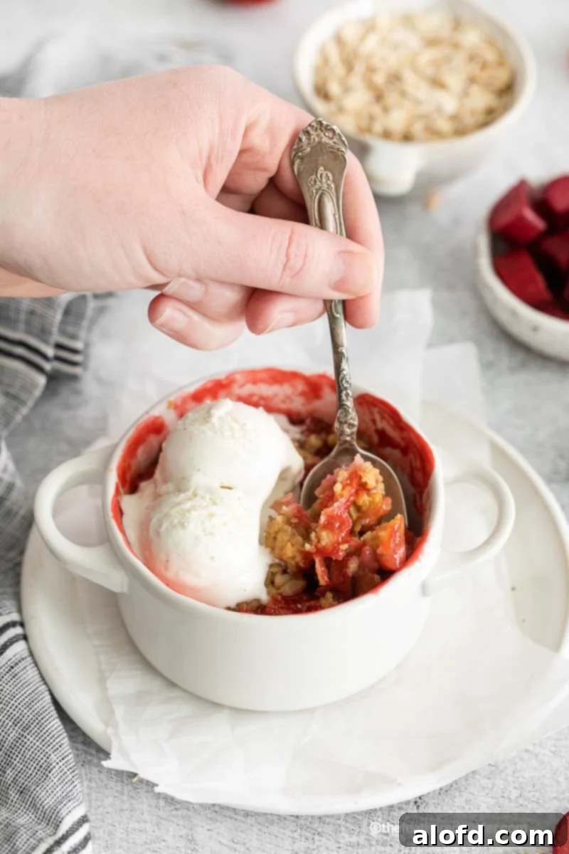 A hand scooping apple strawberry crisp with vanilla ice cream from a ramekin.