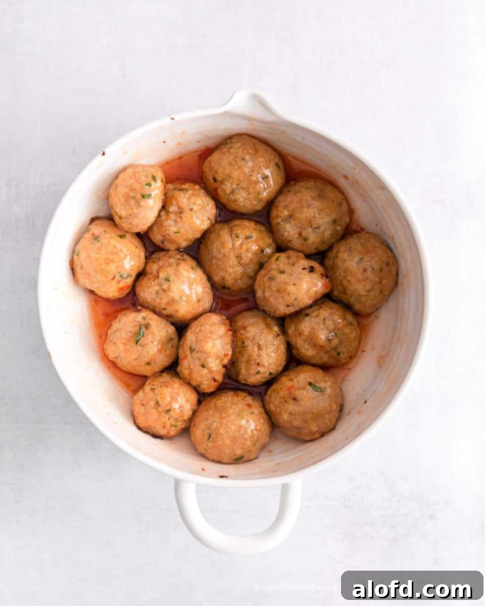 A white mixing bowl with freshly baked sweet chili meatballs being tossed in the vibrant sweet chili sauce, ensuring every meatball is coated.