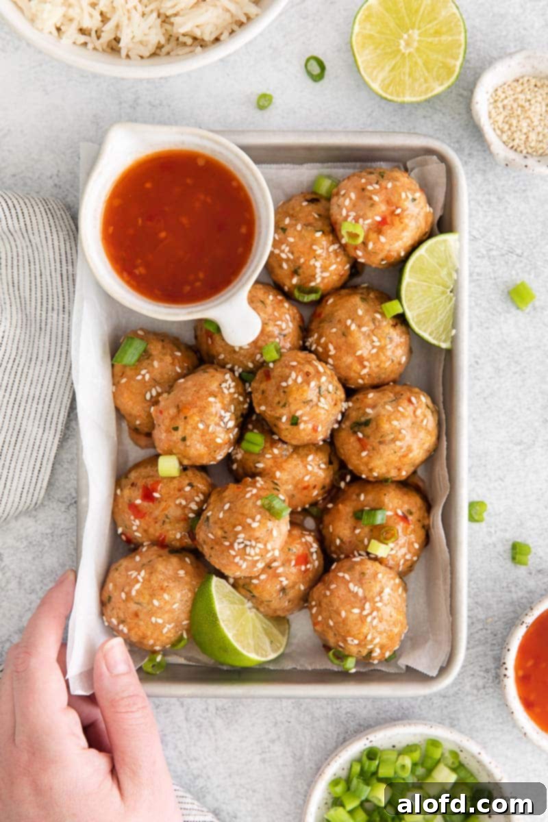 A hand holding a baking sheet showcasing glistening sweet chili meatballs. In the background, there's a gray cloth, bowls of green onion slices, sesame seeds, rice, and an additional bowl of dipping sauce.
