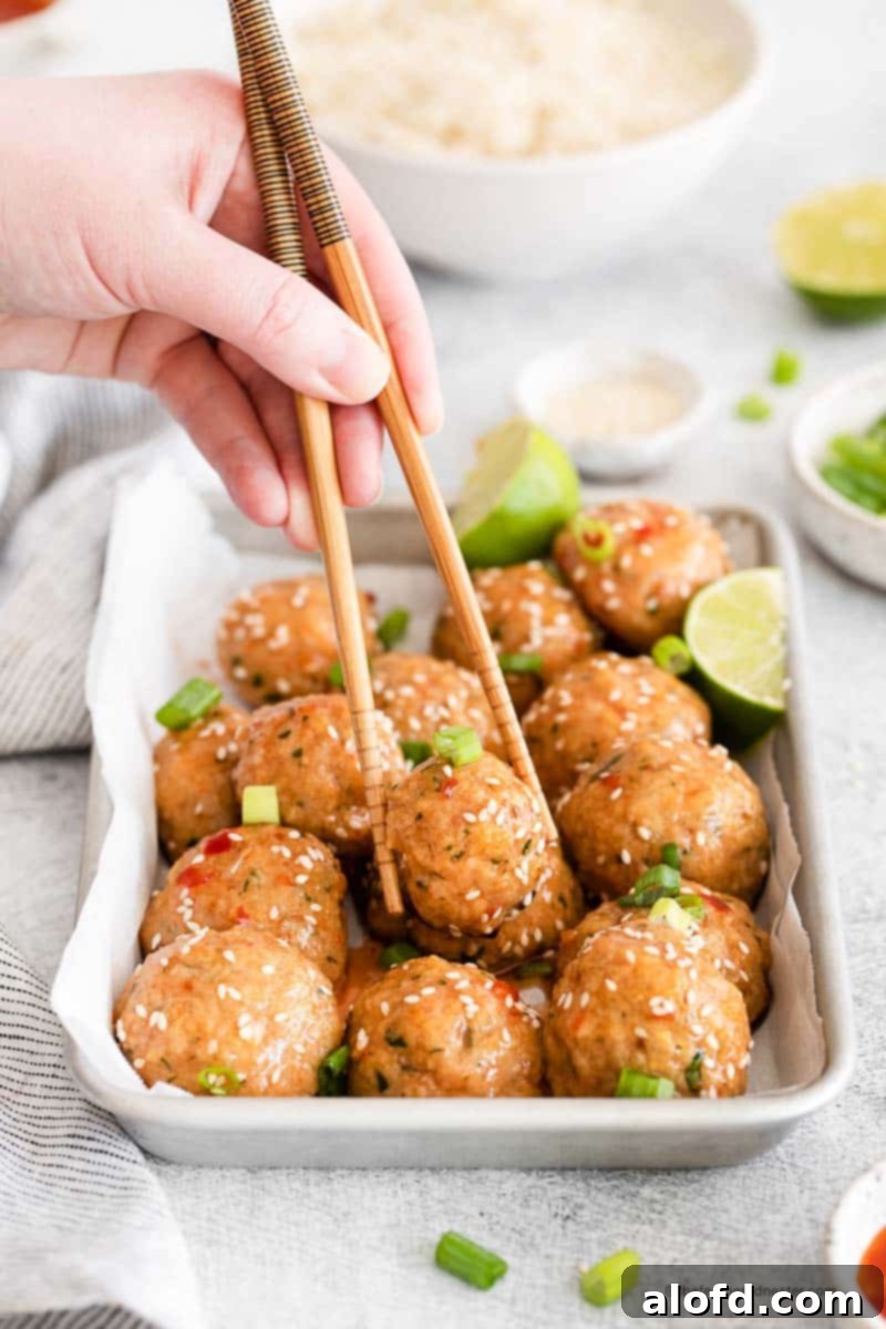 A hand using chopsticks to pick up a sweet chili meatball from a baking sheet, with dipping sauce, lime, sesame seeds, green onions, and rice blurred in the background.