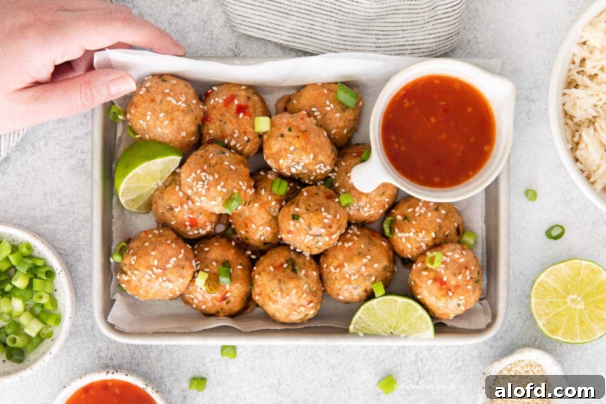 A hand holding a baking sheet with sweet chili meatballs. In the background, there's a gray cloth, bowls of green onion slices, sesame seeds, rice, and an additional bowl of dipping sauce.
