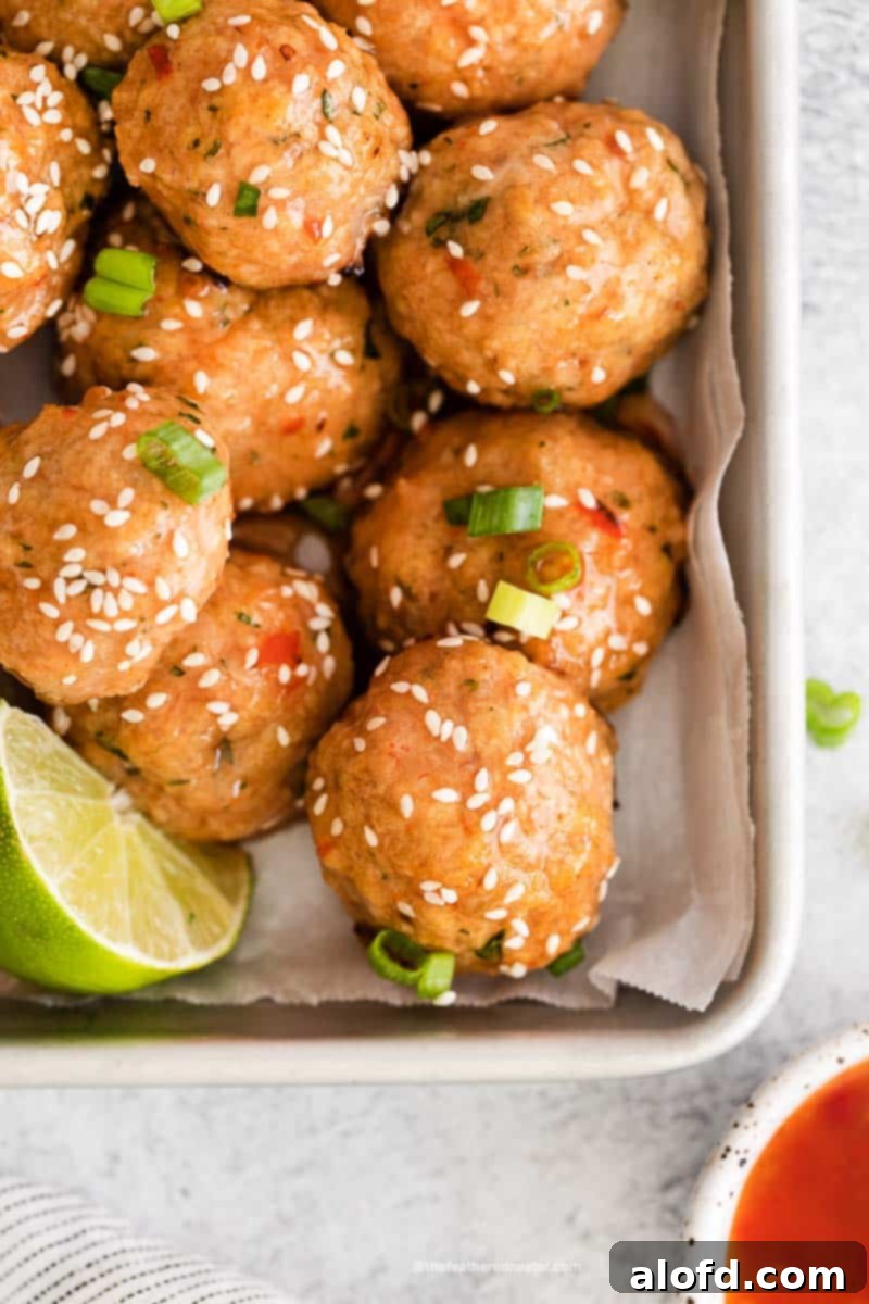A close-up of a baking sheet featuring glazed sweet chili meatballs, accompanied by a vibrant lime slice and a small bowl of dipping sauce, set against a rustic backdrop.