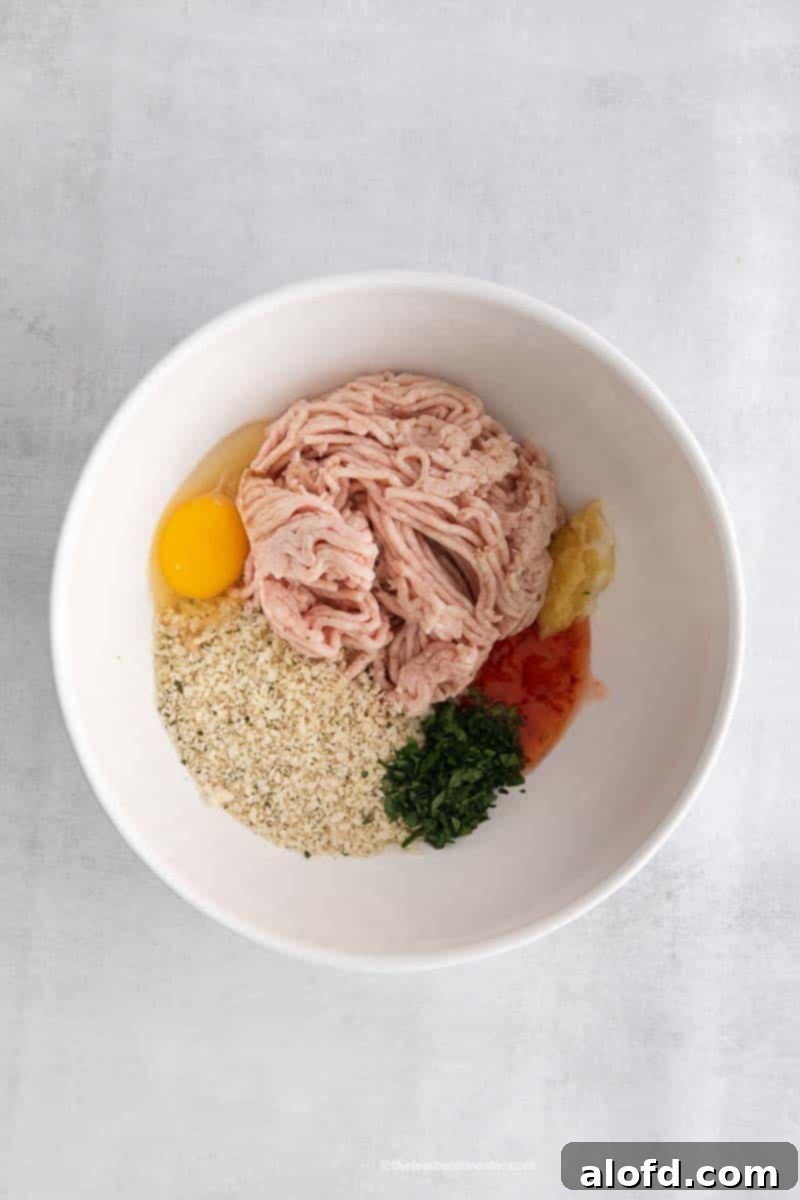 A white mixing bowl filled with raw ground chicken, breadcrumbs, egg, cilantro, and spices, ready to be mixed for sweet chili meatballs.