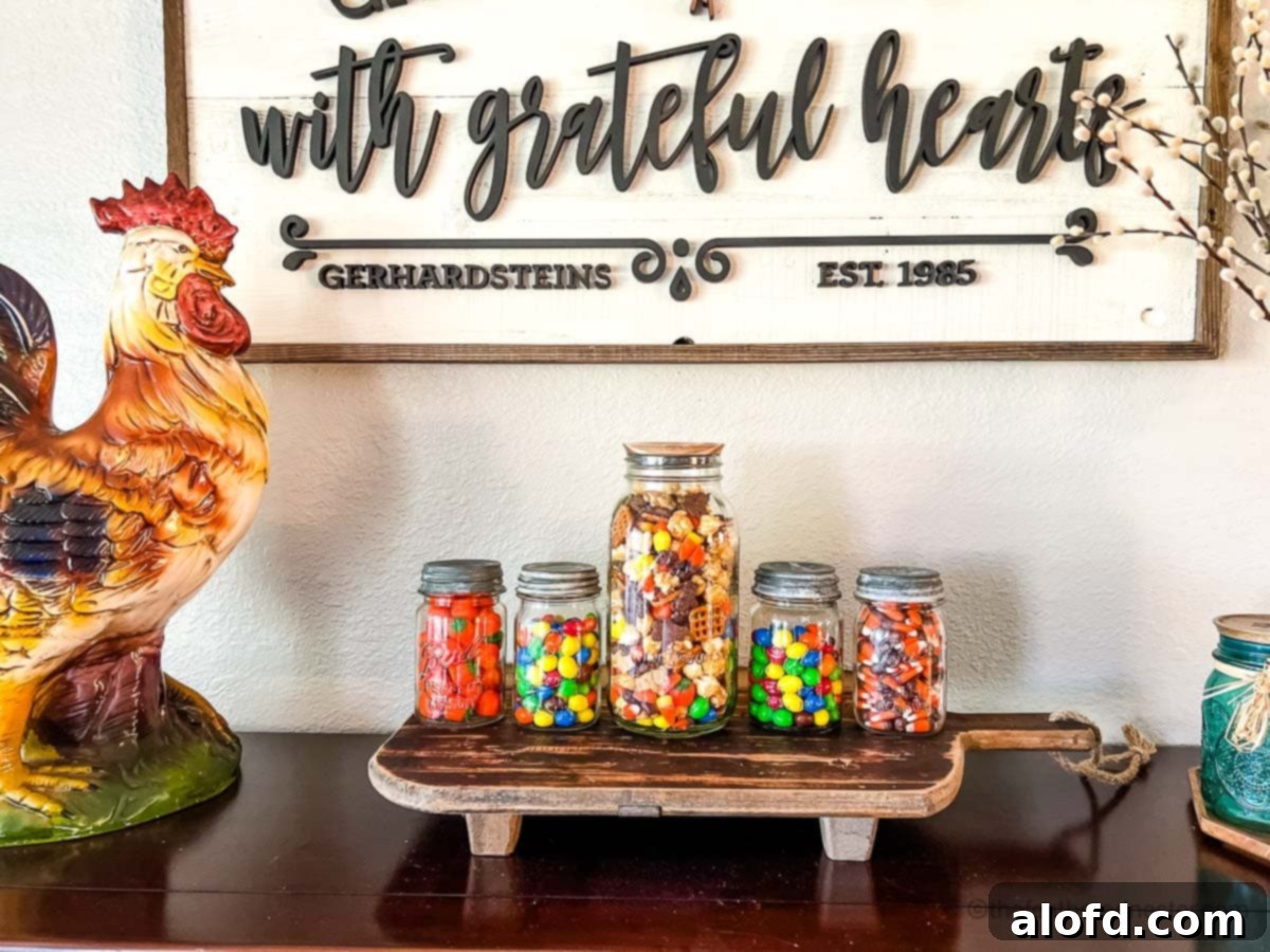 Monster Munch Mix 3 Four small and one large canning jars showcasing a variety of fall and Halloween candies and mixes, elegantly arranged on a rustic wooden board atop a sideboard.