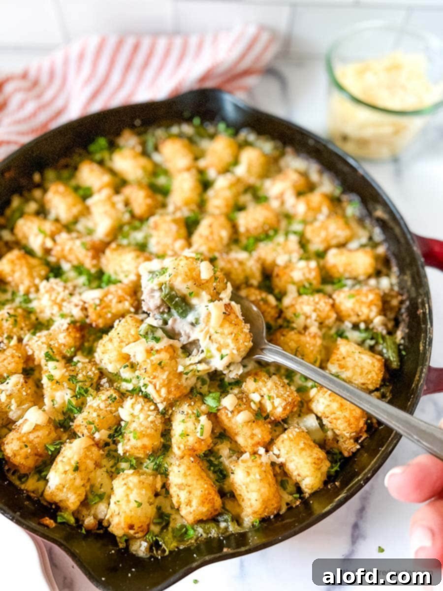 Savory Turkey Tater Bake 2 Turkey Tater Tot Casserole in a cast iron skillet with a hand holding a spoon scooping the turkey tater tots and a red striped cloth, and a jar of shredded cheese in the background.
