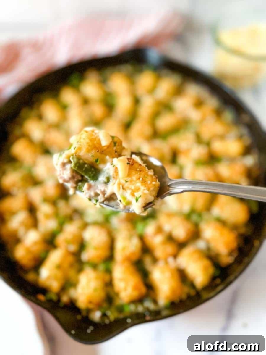 Savory Turkey Tater Bake 11 a spoon with a scoop of turkey tater tot casserole and a skillet full of tater tots, striped red cloth, and a jar of shredded cheese in the background.