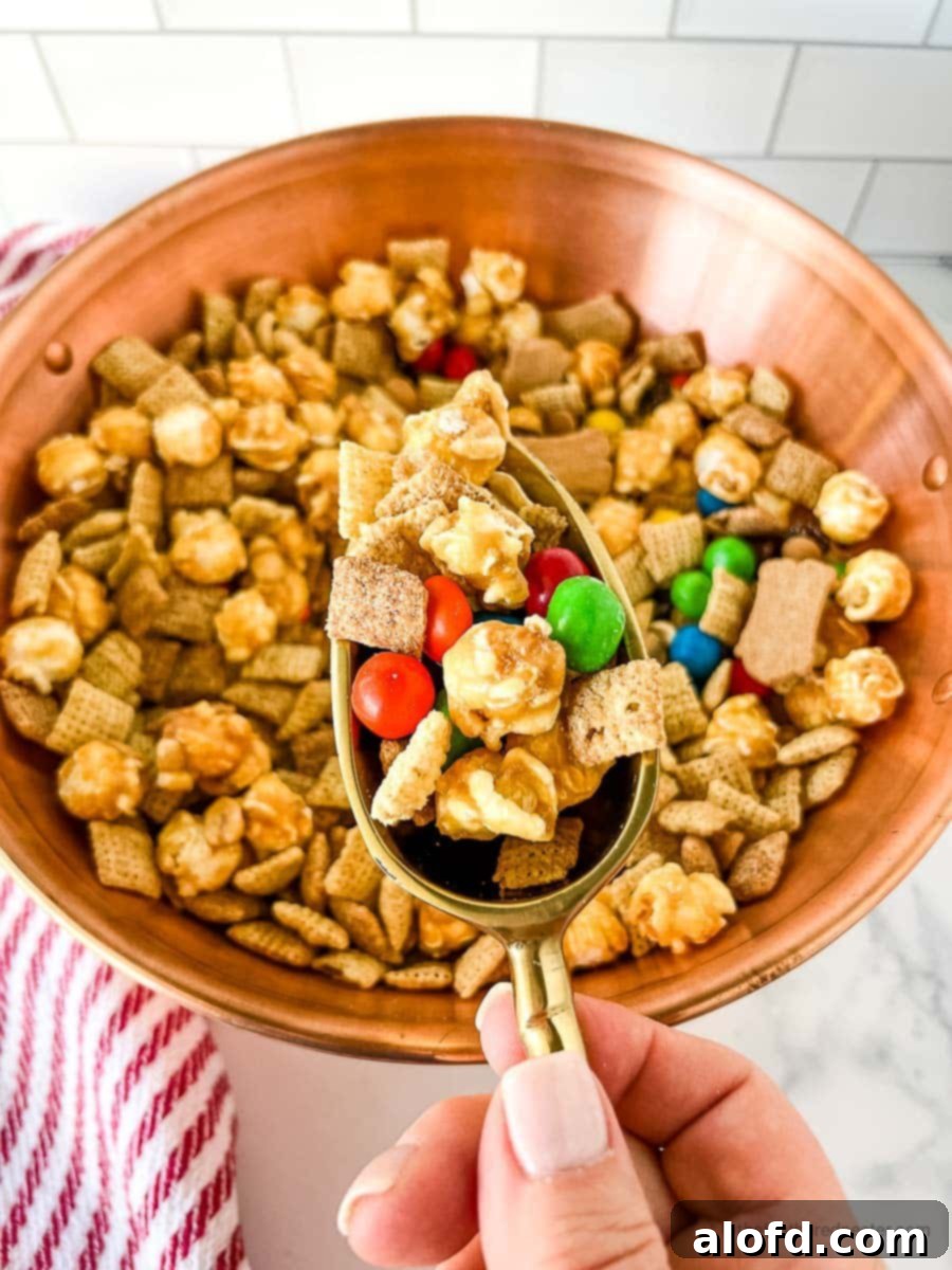 Irresistible M&M Chex Party Snack 3 A hand holding a scoop of delicious Chex party mix with M&M's and various crunchy treats.