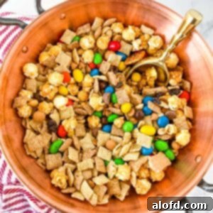 Irresistible M&M Chex Party Snack 8 A close-up photo of a copper bowl with handle, filled with Chex Mix with M&M's and a golden scoop, with a striped kitchen towel in the background.
