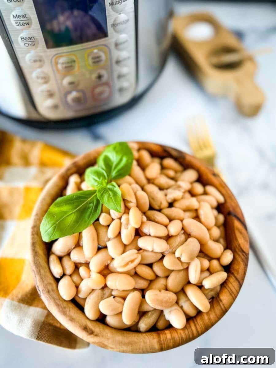 Perfectly Tender Instant Pot Cannellini Beans 2 A wooden bowl of perfectly cooked cannellini beans, garnished with a sprig of fresh basil. A gold and white napkin and an Instant Pot are visible in the soft-focus background, suggesting a home kitchen setting.